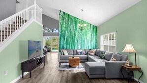 Ein geräumiges Wohnzimmer mit einer grauen Eckcouch, einem Holztisch und einem Fernseher auf einem schwarzen Standfuß. Eine Treppe mit weißen Geländern ist links sichtbar, und ein grüner Vorhang hängt im Hintergrund.