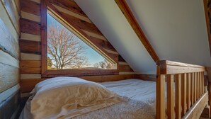 A bedroom with a sloped ceiling, wooden beams, a window, and a fluffy white bedspread. - Historical Paris Cabin on Farm w/ Fire Pit! (Paris)