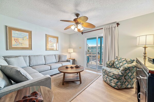 A living room with a sofa, armchair, coffee table, and ceiling fan. There are framed pictures on the wall and a view of the outside through a window.