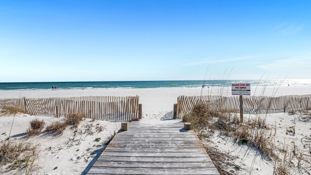 Une promenade en bois mène à une plage de sable, et un panneau indique qu’il est interdit de s’approcher des dunes.