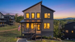 A modern multi-story house with a balcony, large windows, and an American flag. - Sleek 4BR Cabin with Hot Tub, Theater & Arcade | Golf & Waterpark Nearby! (Sevierville)