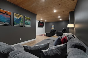 A modern living room with a grey couch, a black recliner, and a wooden ceiling. There are posters on the wall and a lamp on the right. - Sleek 4BR Cabin with Hot Tub, Theater & Arcade | Golf & Waterpark Nearby! (Sevierville)