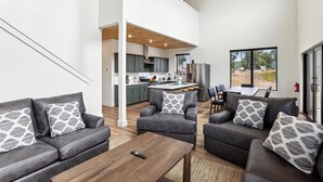 Una sala de estar moderna con sofás grises y cojines estampados, una mesa de centro de madera, y vistas a la cocina y al área exterior.