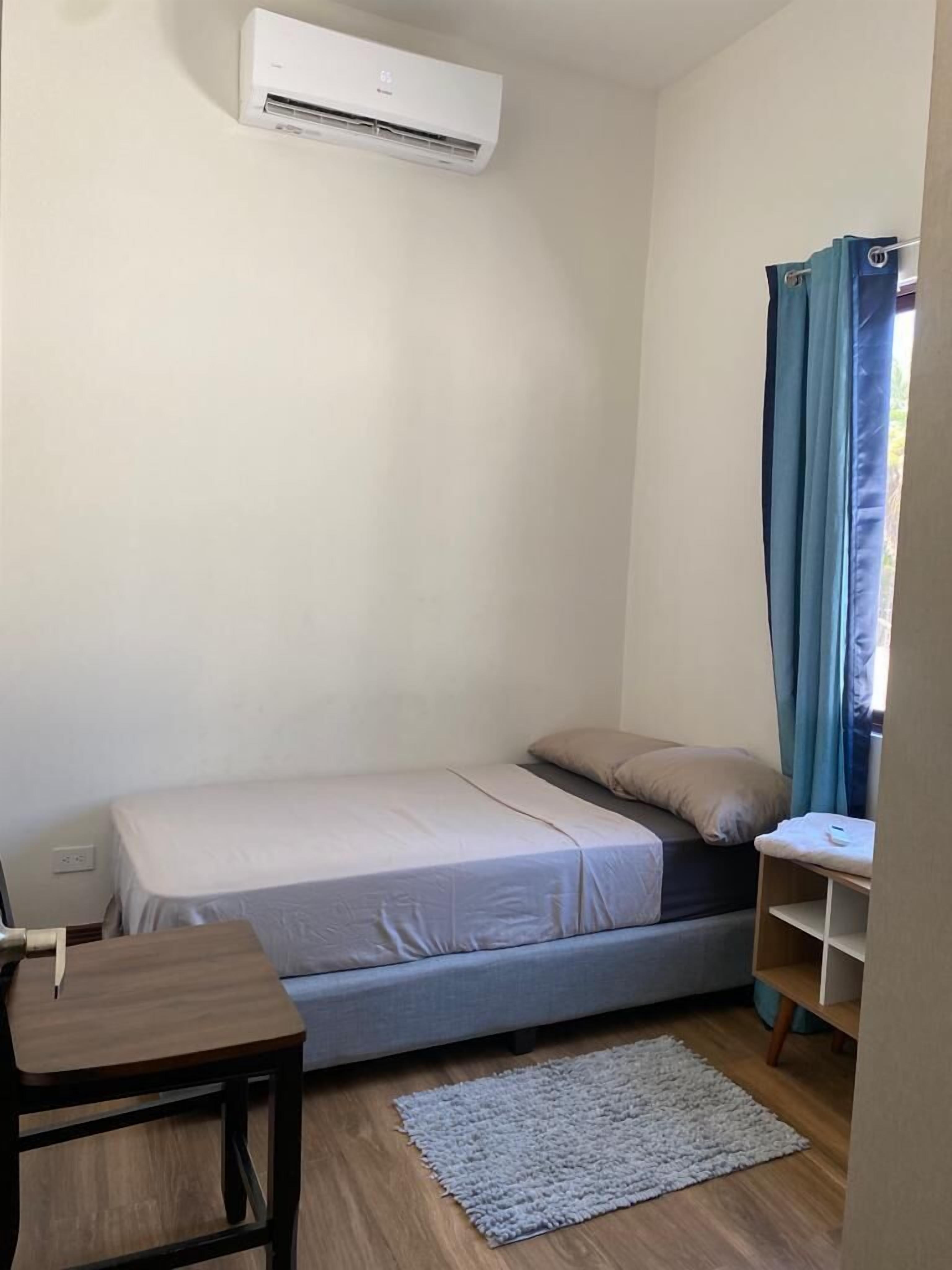 Une petite pièce, de taille modeste, comprenant un lit simple, une table en bois, une petite étagère et un tapis gris.