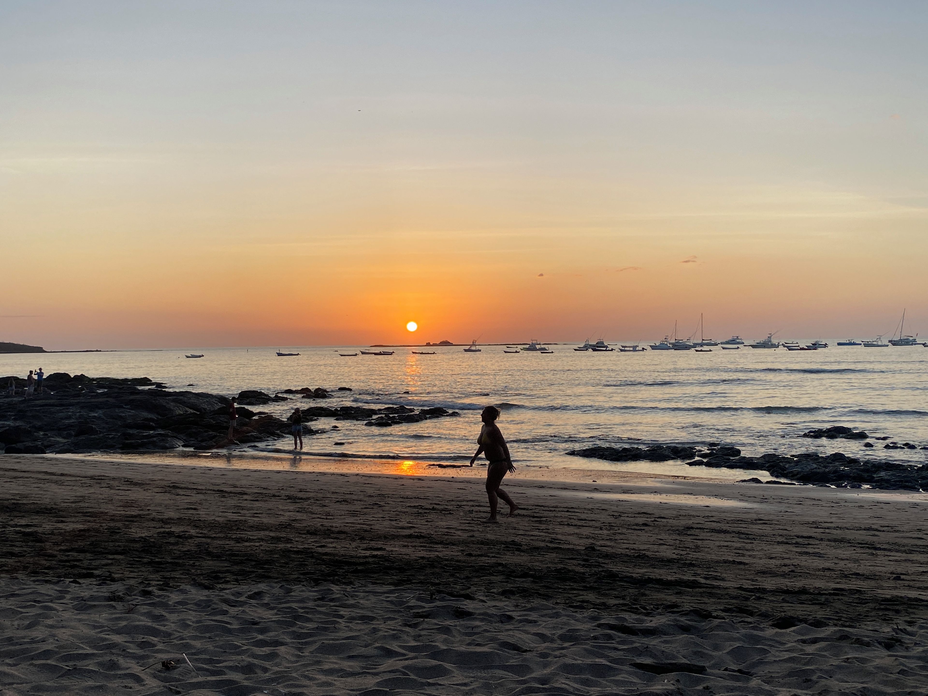 Eine Strandansicht mit einer Person, die am Ufer entlanggeht, Booten im Wasser und einem Sonnenuntergang im Hintergrund.