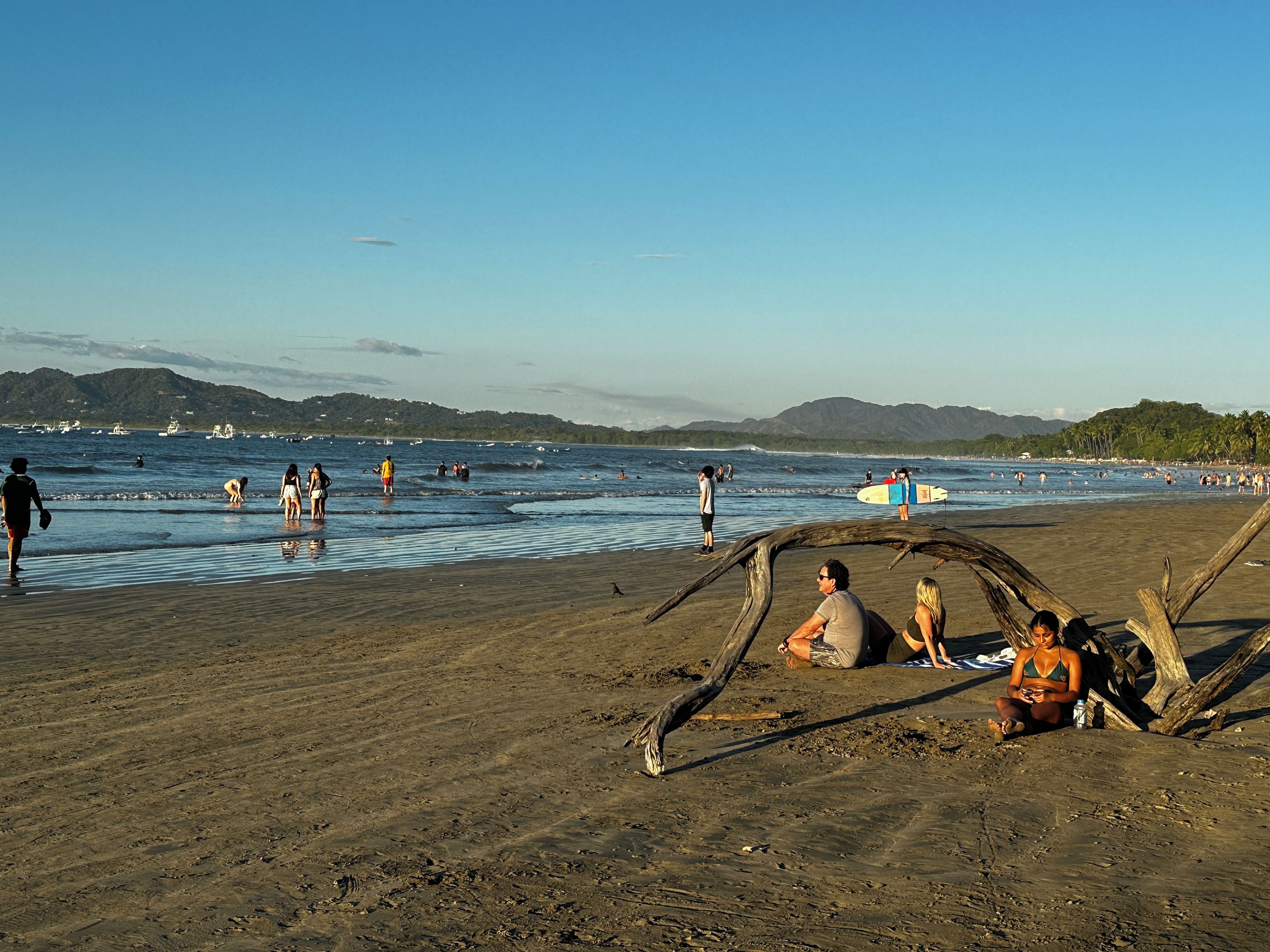 Una playa con gente, una tabla de surf y barcos en el agua.