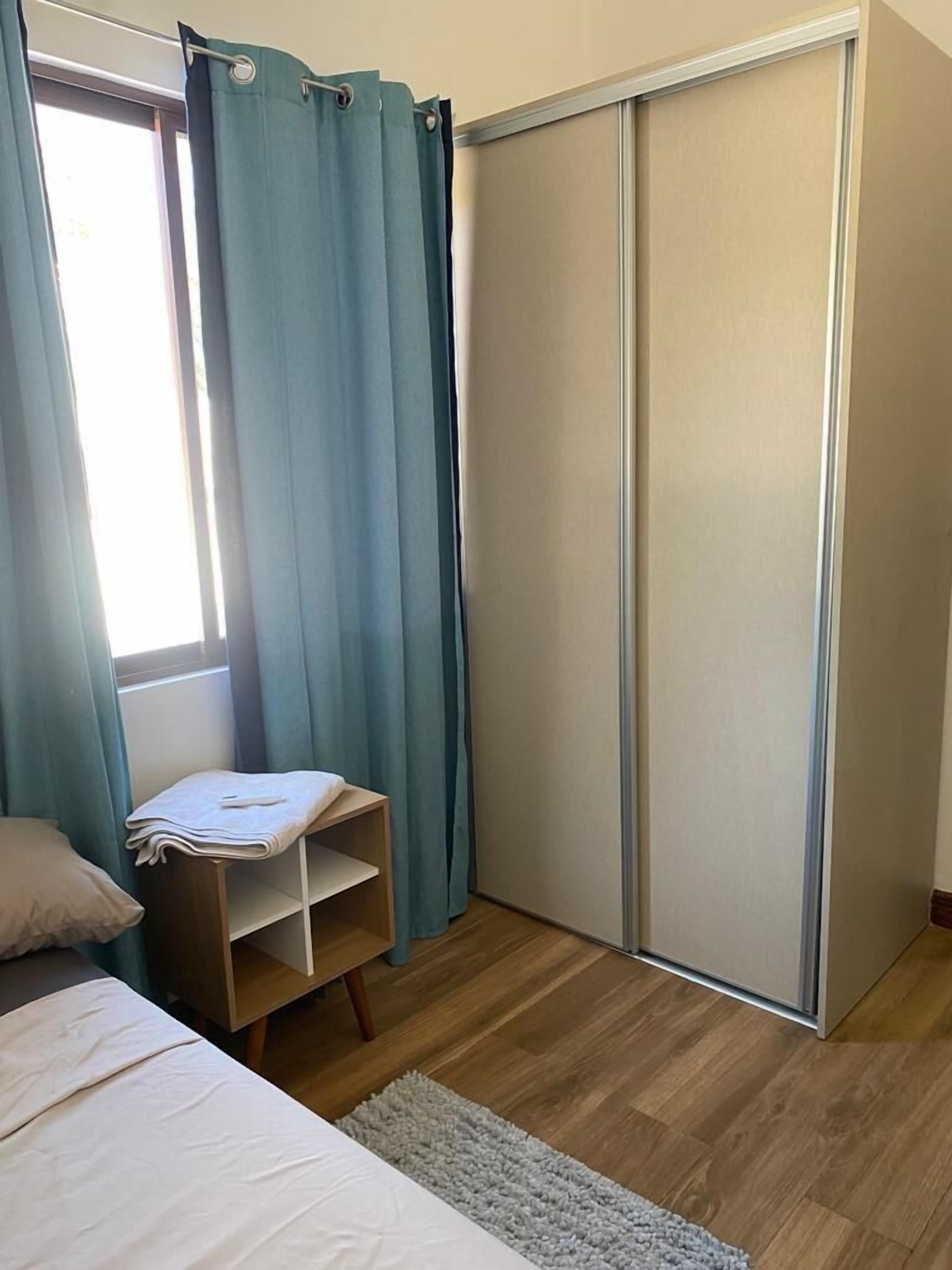 Quarto com cama, uma pequena prateleira, guarda-roupa e janela com cortinas.
