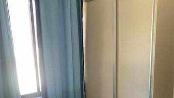 Un dormitorio con cama, una pequeña estantería, un armario y una ventana con cortinas.