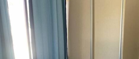 Quarto com cama, uma pequena prateleira, guarda-roupa e janela com cortinas.