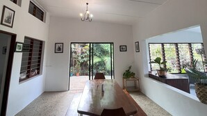 Una habitación amplia con una larga mesa de madera y bancos, una araña y ventanas grandes con vistas a la vegetación.