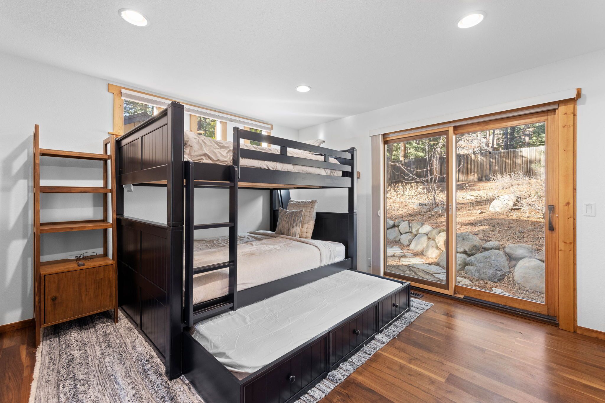 Un lit superposé avec une échelle en bois et un petit placard, une porte-fenêtre coulissante donnant sur un espace extérieur avec des rochers et des arbres, et un plancher en bois.