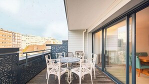 A balcony with a table and chairs, a glass door leading to an interior room, and a view of buildings in the background.