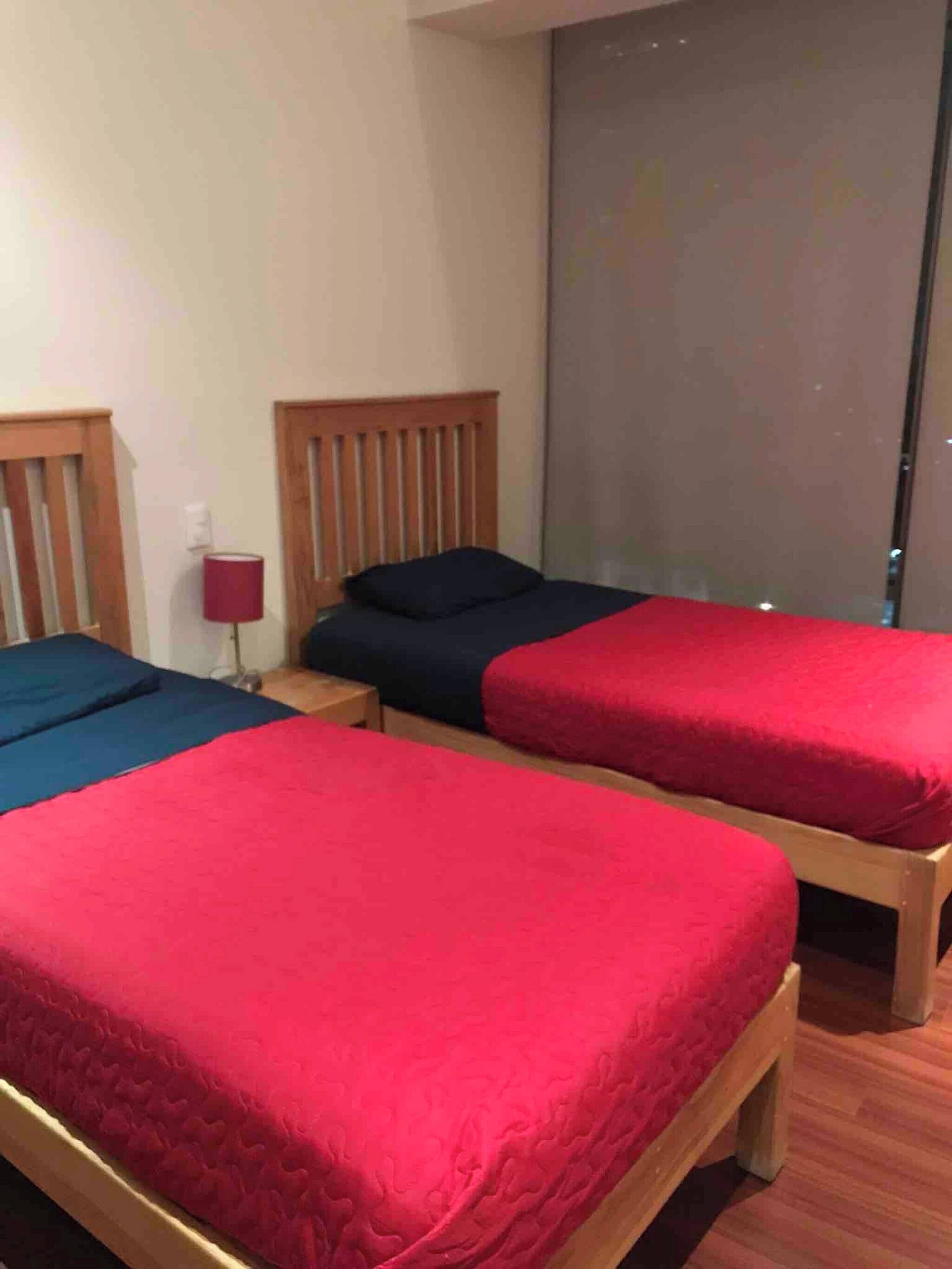 Una habitación con tres camas individuales, cada una con cabecera de madera, colcha roja y mesita de noche con lámpara.