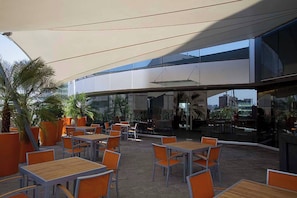An outdoor seating area with tables and chairs, large potted plants, and a modern building in the background.
