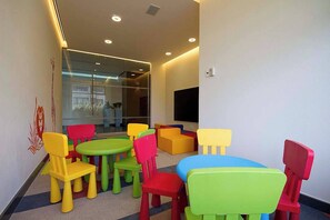 Un área de juegos para niños con sillas y mesas de plástico de colores, un ventanal grande y un televisor de pantalla plana.