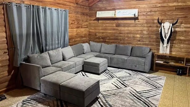 Interno di una baita in legno con un divano componibile, poggiapiedi, pareti di legno, una ventola a soffitto e un teschio di mucca decorativo.