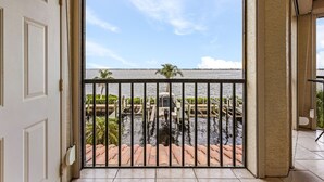 A view through a balcony door of a water body with a dock and palm trees. - RIVERFRONT VIEW, Stunning Sunrises, Community Pool - Condo Waterfront Sunrise - Roelens (Cape Coral)