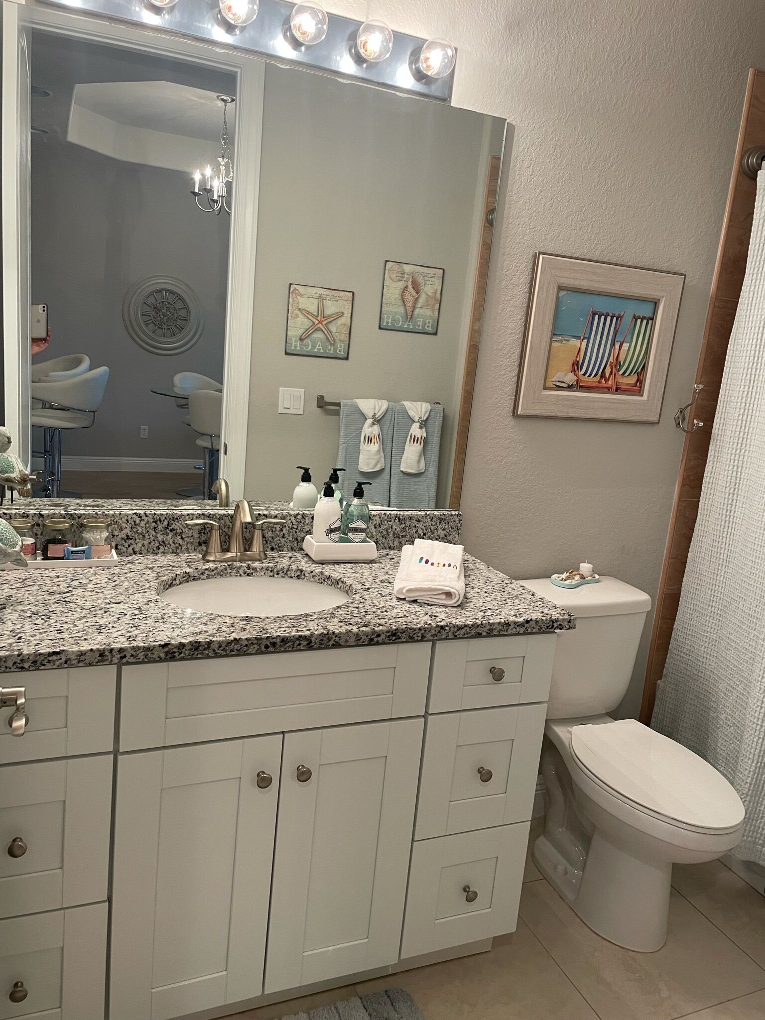 A bathroom with a double sink vanity, a large mirror with lights, a toilet, and a framed beach picture on the wall.