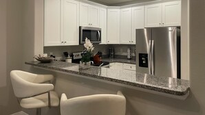 Una cocina moderna con armarios blancos, un refrigerador de acero inoxidable, un microondas y una encimera de granito. Hay dos taburetes blancos y un florero con flores sobre la encimera.