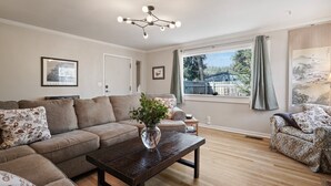 Uma sala de estar com sofá seccional, uma mesa de centro com um vaso, um lustre, uma janela grande com cortinas e um quadro na parede.