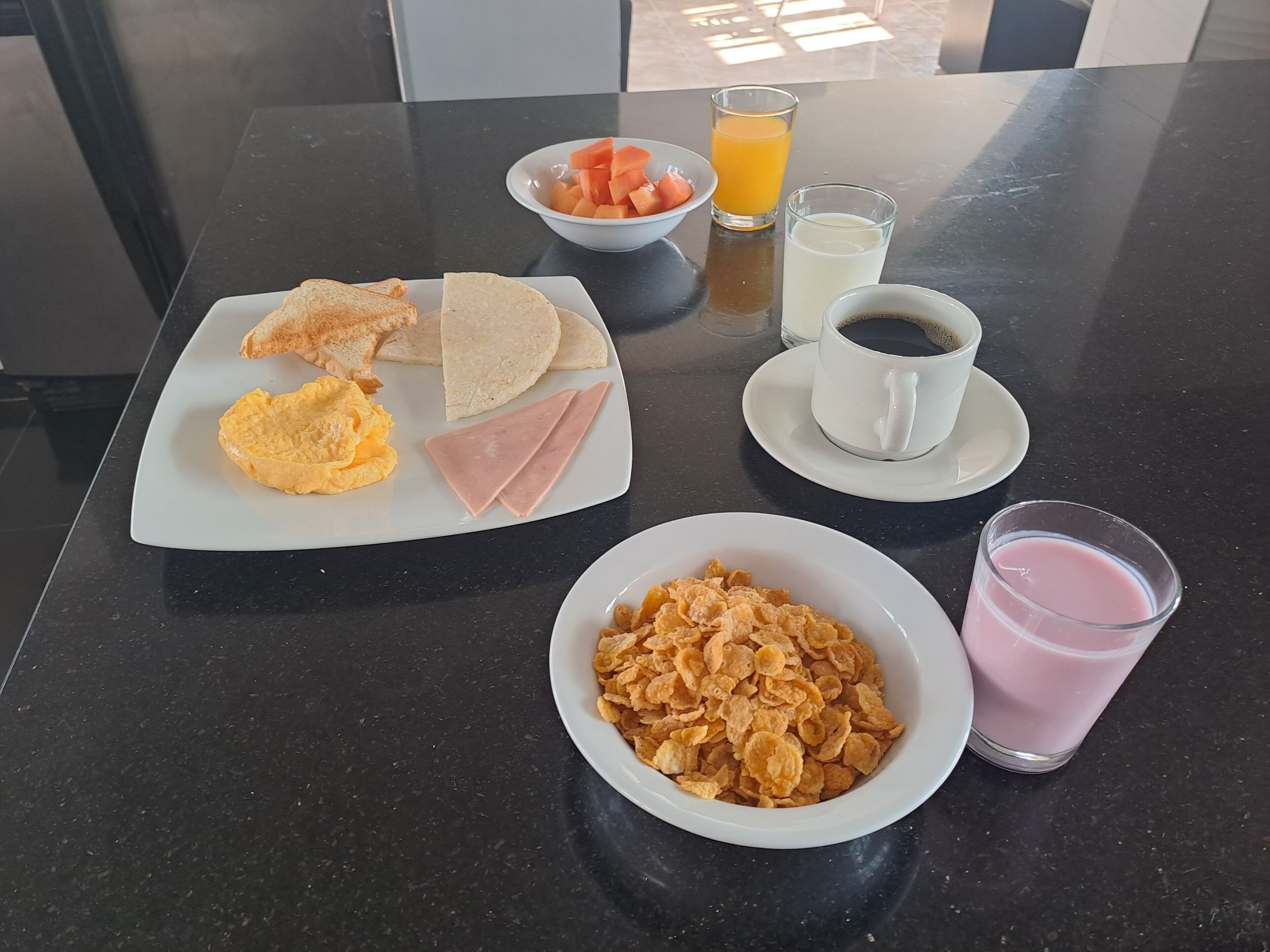 Sur la table, on trouve divers aliments pour le petit-déjeuner : des toasts, des œufs, du jambon, des céréales, du jus de fruits et du café.