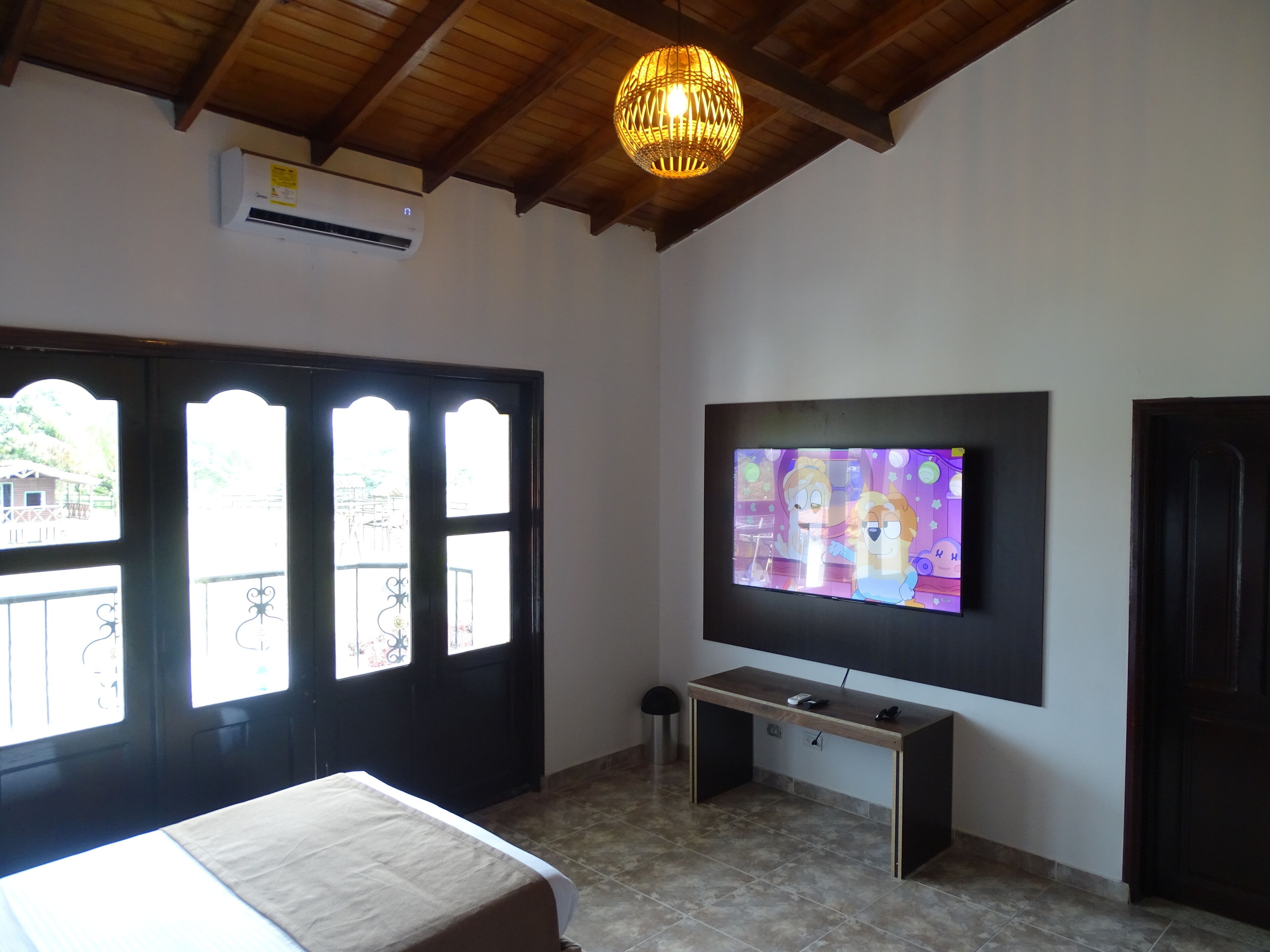A room with a bed, a TV mounted on the wall, a desk, and a window with a view of the outside.