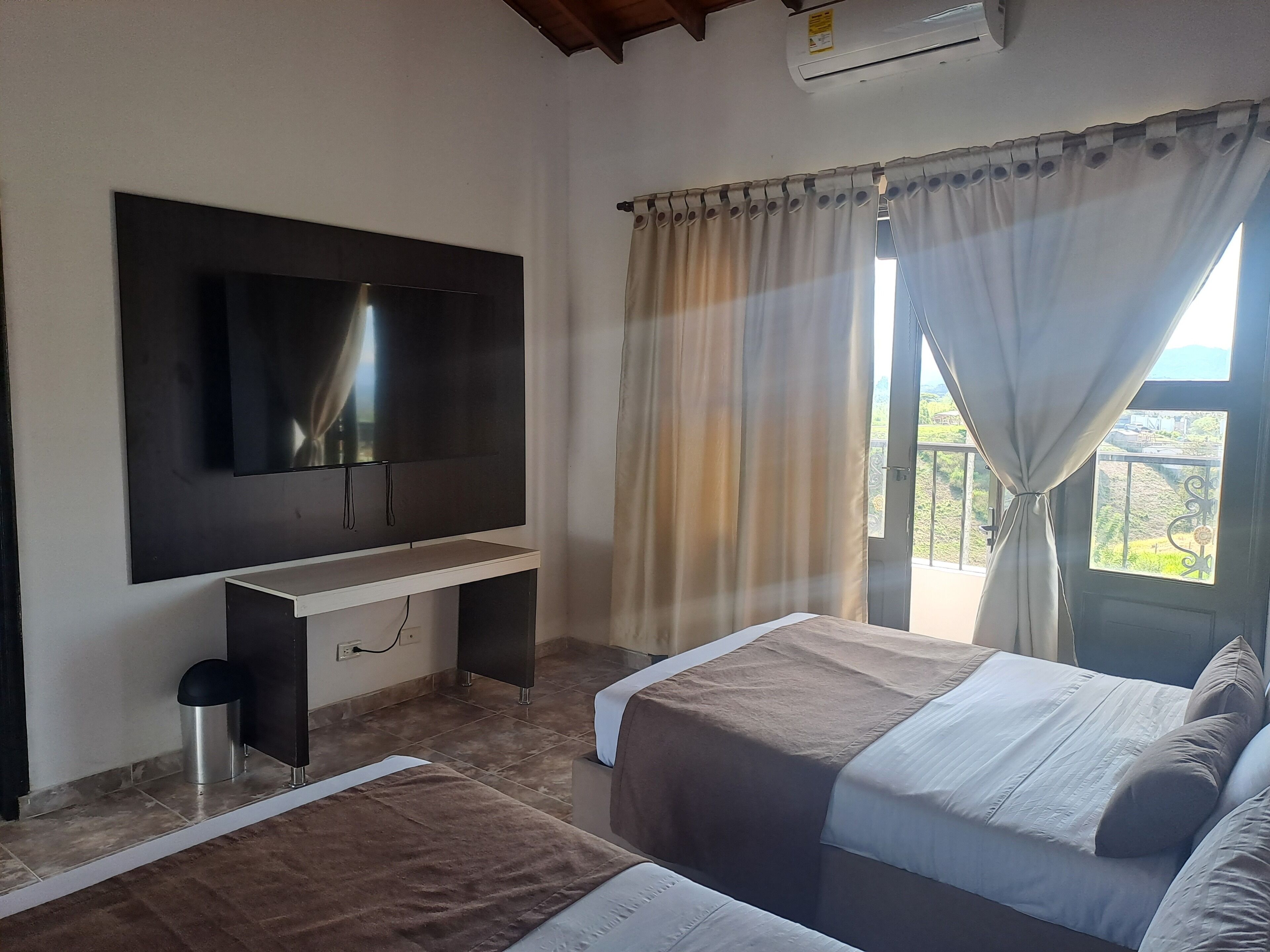Une chambre à coucher avec un téléviseur fixé au mur, un lit avec une tête de lit, une table de chevet et une fenêtre avec des rideaux.