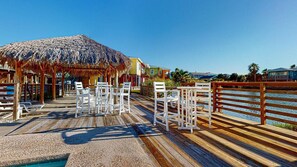 Una terraza de madera con sillas blancas y un paraguas de paja, con vistas a una piscina y propiedades frente a la playa.