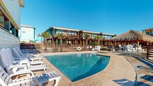 A pool area with lounge chairs, a thatched umbrella, and a building with multiple balconies. - OV17 Shared Pool, Pet Friendly, Golf Cart Access, Ping Pong Table (Port Aransas)