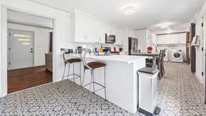 A modern kitchen with white cabinets, a central island with stools, a microwave, a refrigerator, and a washing machine. - East Point Retreat Near ATL Airport (East Point)
