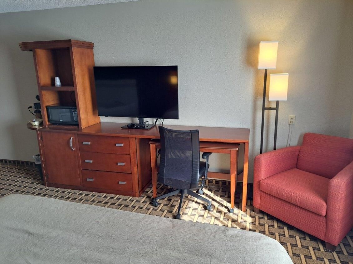 A hotel room with a bed, a desk with a TV, a red chair, a lamp, and a wooden cabinet with drawers.