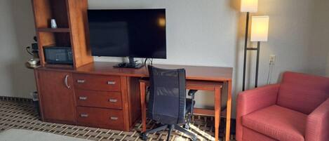 A hotel room with a bed, a desk with a TV, a red chair, a lamp, and a wooden cabinet with drawers.