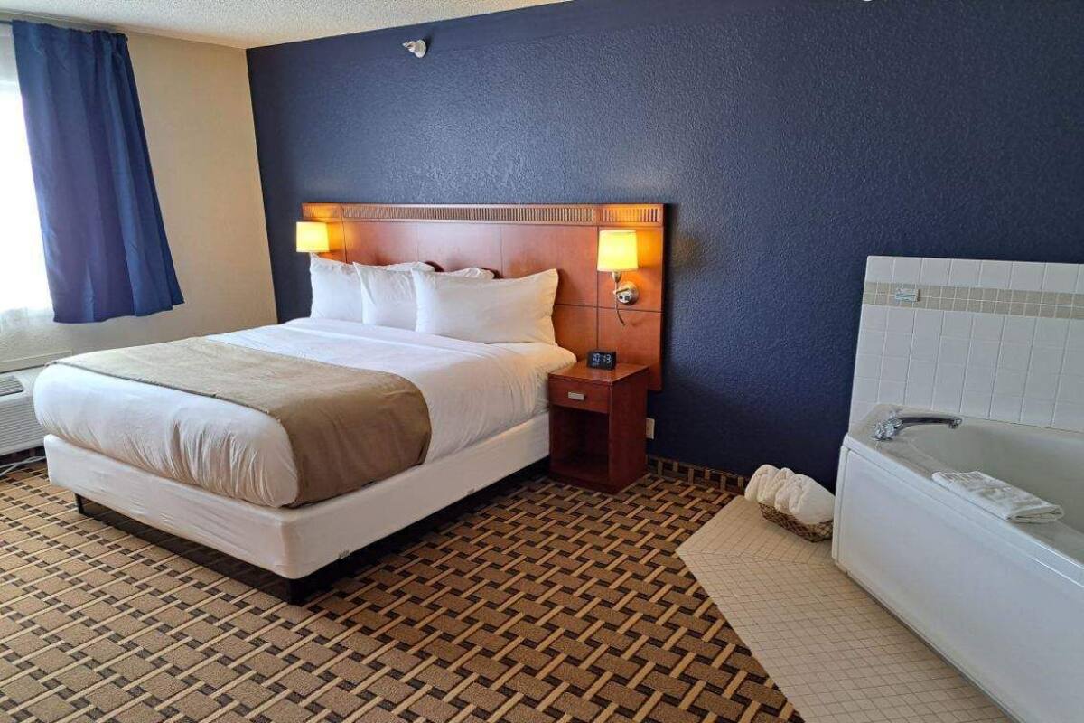 A hotel room with a large bed, a bedside table with a lamp, a bathroom with a bathtub, and a patterned floor.