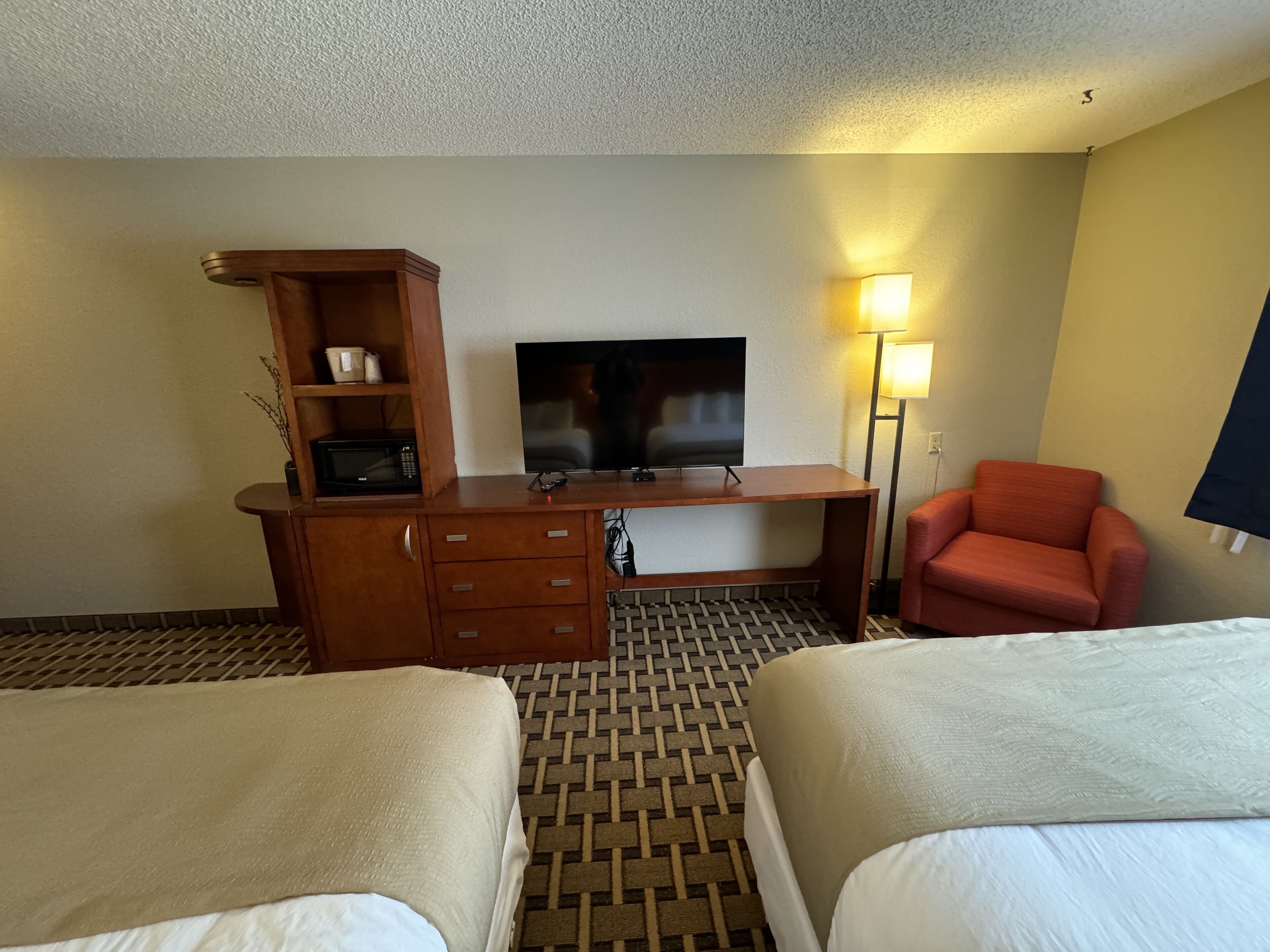 Una habitación de hotel con dos camas, un escritorio con televisor, microondas, lámpara y una silla roja.