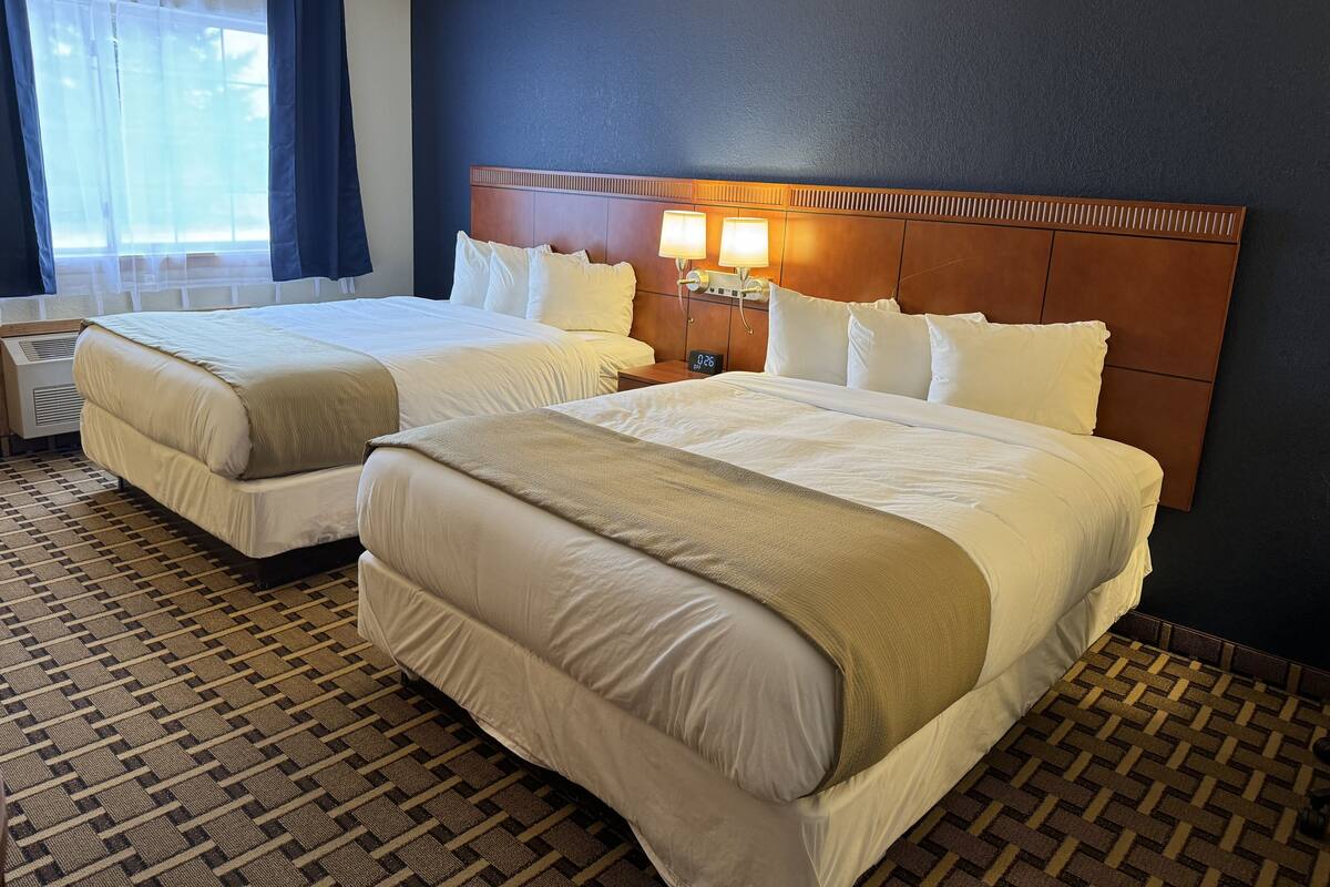 A hotel room with two beds, a large window with curtains, a headboard with a clock, and a patterned carpet.