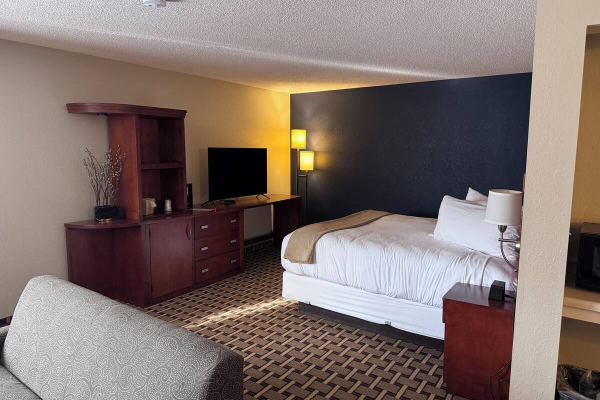 A hotel room with a bed, a desk with a flat-screen TV, a lamp, a dresser, and a patterned chair.