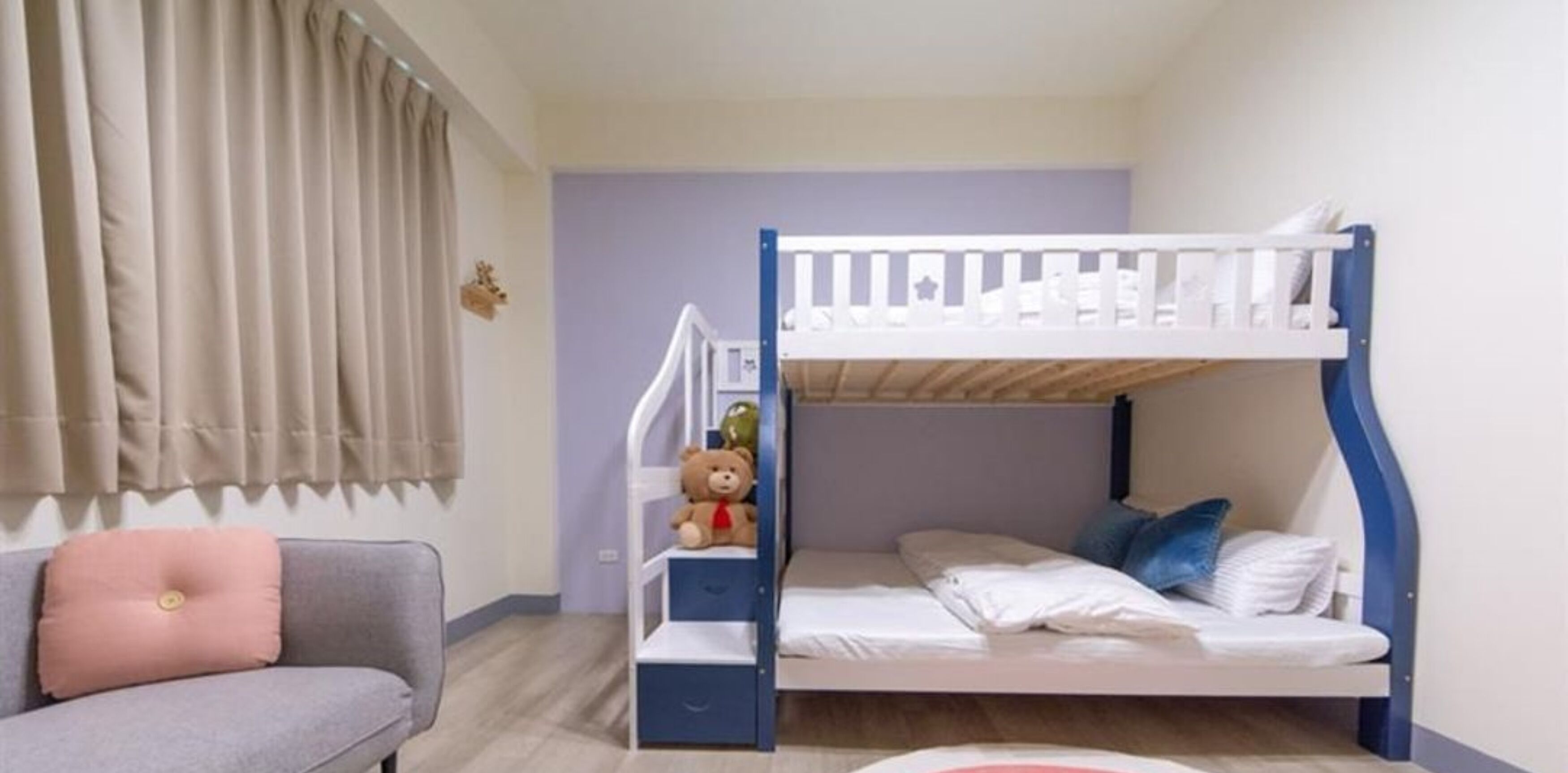 A bunk bed with a white frame and blue accents, a grey sofa with a pink cushion, and a teddy bear on the bed.