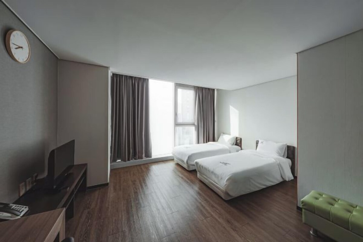 Habitación de hotel con dos camas, un escritorio, un televisor y un ventanal grande con cortinas.