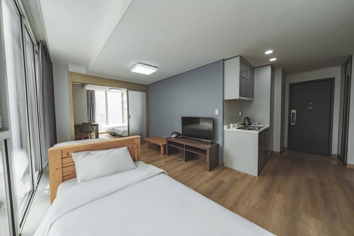 Ein Hotelzimmer mit Bett, Schreibtisch, Fernseher, einer kleinen Küchenzeile und einer Tür.