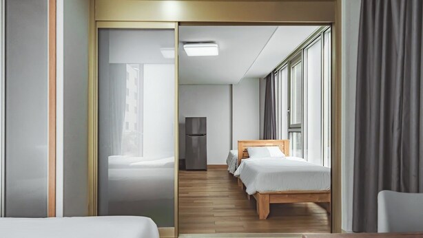 Une chambre d’hôtel moderne avec un plancher en bois, deux lits simples, un réfrigérateur et de grandes fenêtres équipées de rideaux.