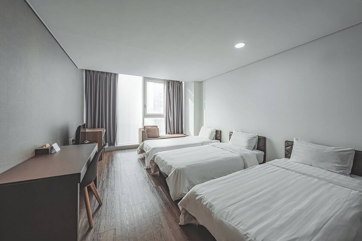 Ein Hotelzimmer mit zwei Betten, einem Schreibtisch und einem Stuhl. Es gibt ein Fenster mit Vorhängen und Blick auf die Stadt.