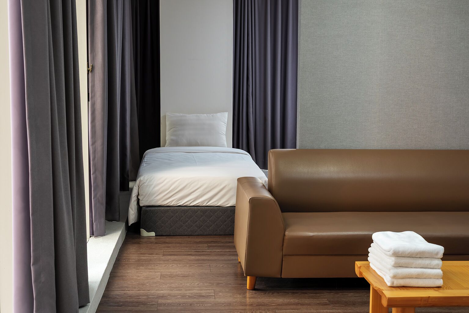 Hotellihuone, jossa on sänky, ruskea sohva, puinen pöytä, jossa on pyyhkeitä, ja tummat verhot.