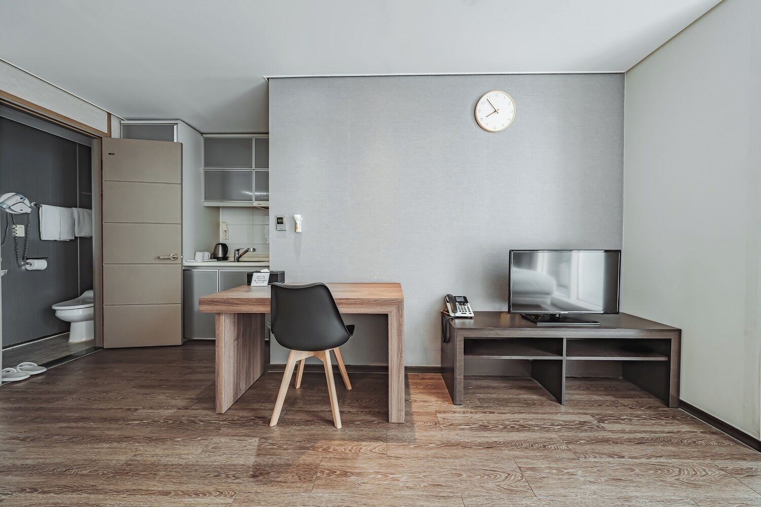 A compact living space with a kitchenette, a wooden desk, a black chair, a TV on a stand, and a clock on the wall.