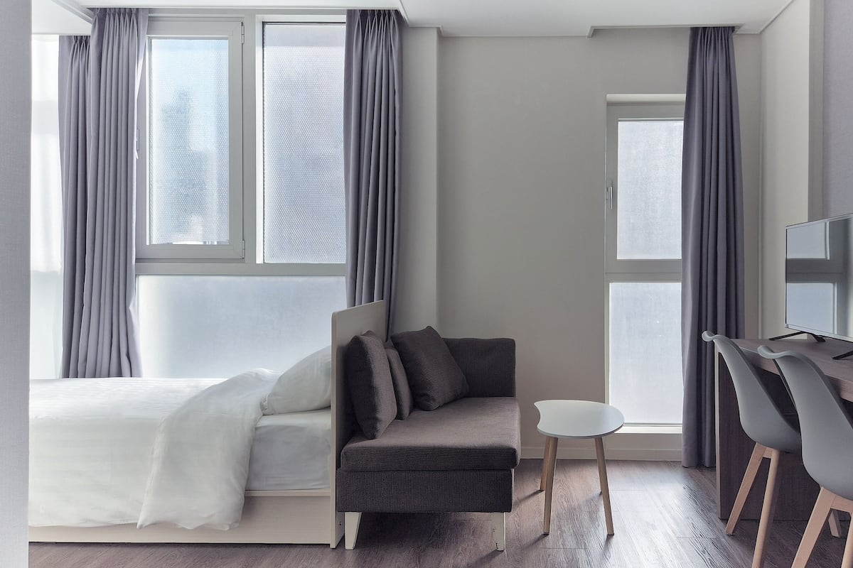 Ein modernes Hotelzimmer mit einem Bett, einer Couch, einem Schreibtisch mit Computer und zwei mit Vorhängen versehenen Fenstern.