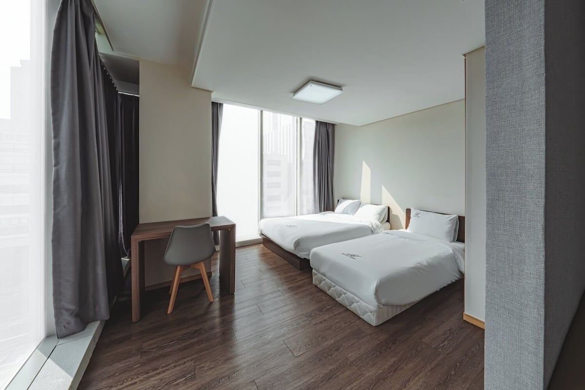 Ein modernes Hotelzimmer mit Schreibtisch aus Holz, zwei Betten und einem großen Fenster mit Vorhängen.