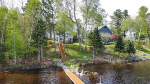A lakeside property with a wooden dock extending into the water, surrounded by trees and a house in the background.