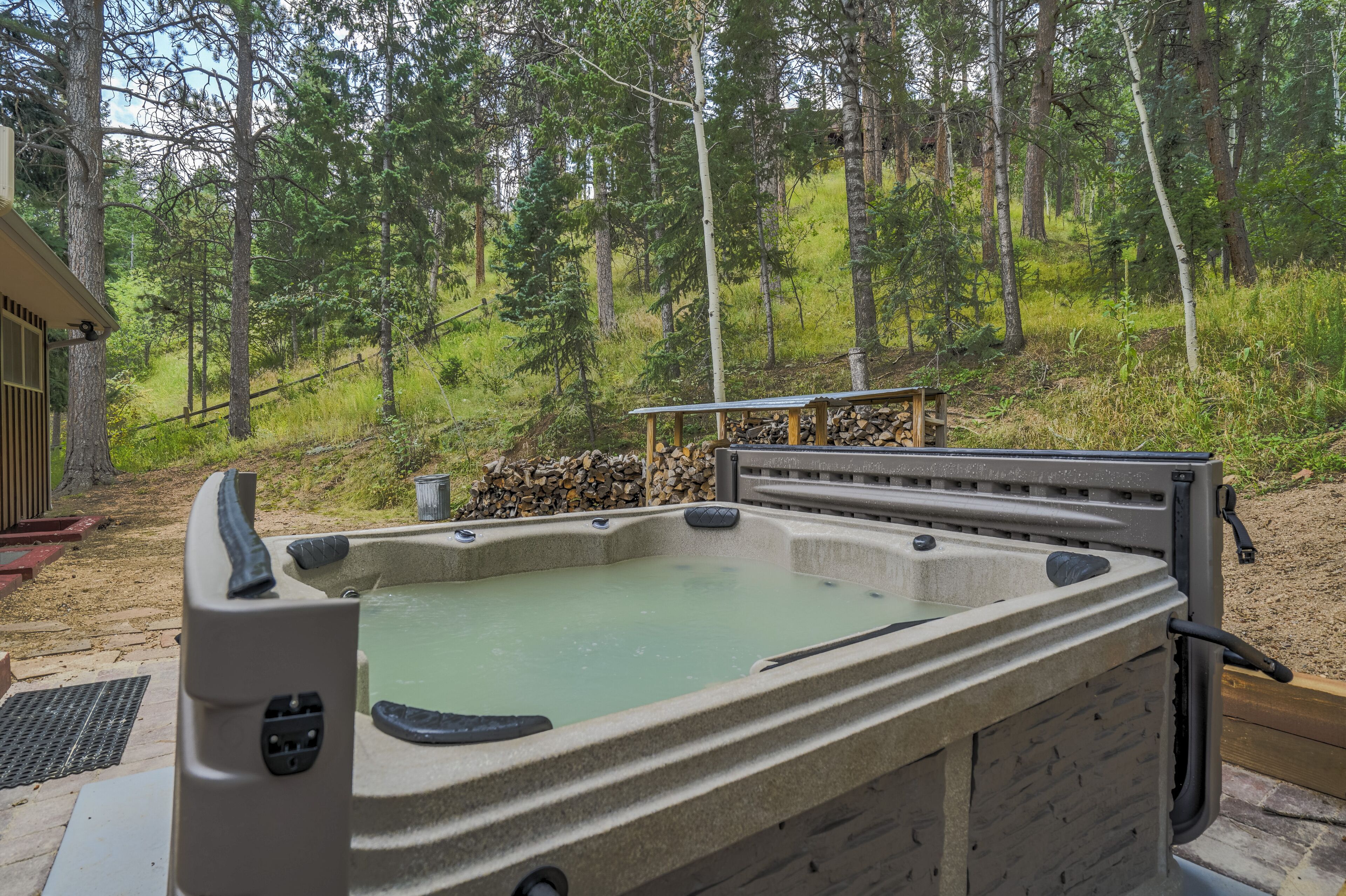 A hot tub in a forested area with a wooden deck and a stack of firewood nearby.