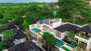 Una villa moderna con piscina, rodeada de exuberante vegetación y otras edificaciones residenciales.