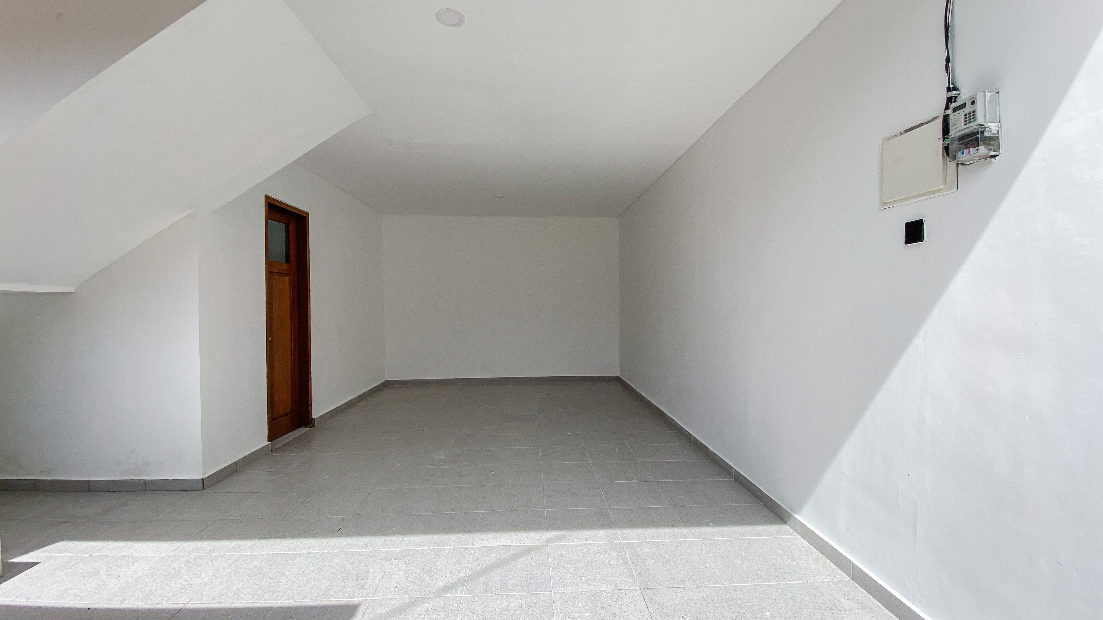 A spacious, empty room with a sloped ceiling, a wooden door, and a wall-mounted electrical box.
