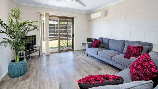 A modern living room with a grey sofa, red and black patterned cushions, a large potted plant, and a ceiling fan. - Gold Rush Apartments (Monkland)